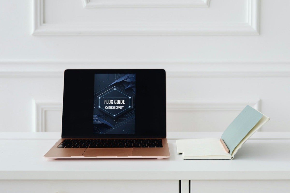 A rose gold laptop sits open on a white desk, displaying a digital course cover titled 'FLUX GUIDE CYBERSECURITY'. To the right of the laptop is an open notebook with a light blue cover.