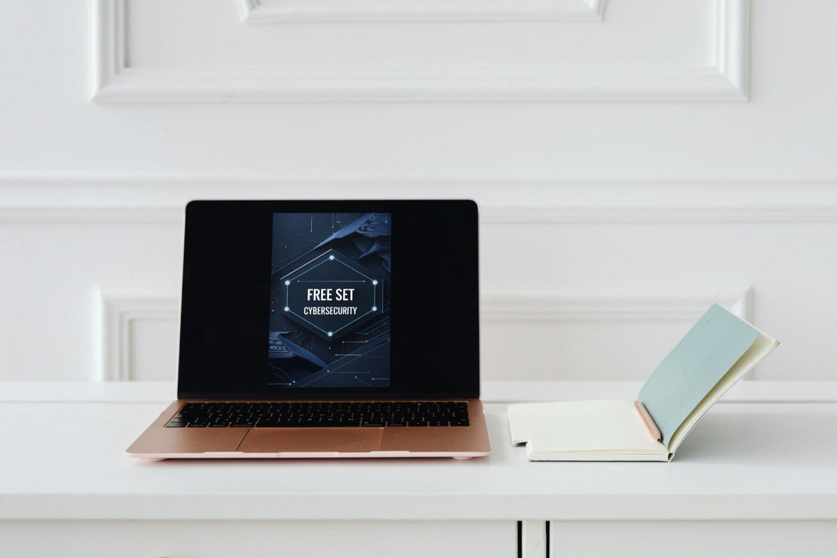 A rose gold laptop sits open on a white desk, displaying a digital graphic with the text 'FREE SET CYBERSECURITY'. To the right of the laptop is an open notebook with a light blue cover.